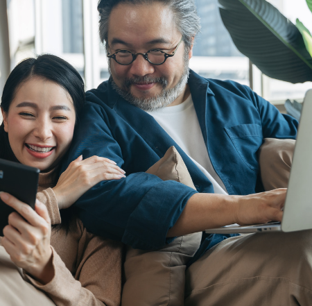 امرأة مبتسمة تعرض هاتفها على رجل مسن مبتسم بينما يكتب هو على حاسوب محمول